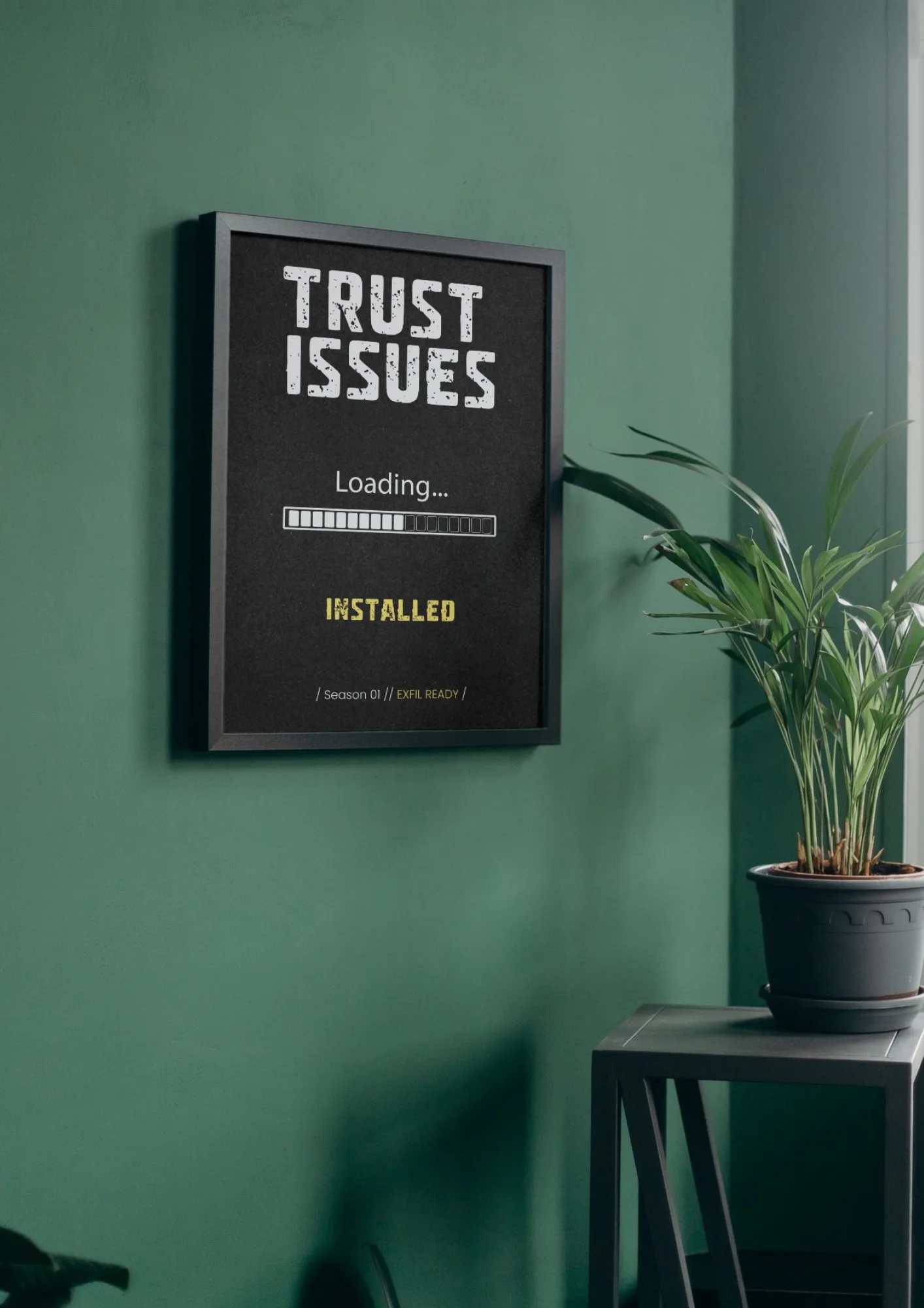 Plakát ARC Raiders TRUST ISSUES v černém rámu na zelené stěně vedle pokojové rostliny