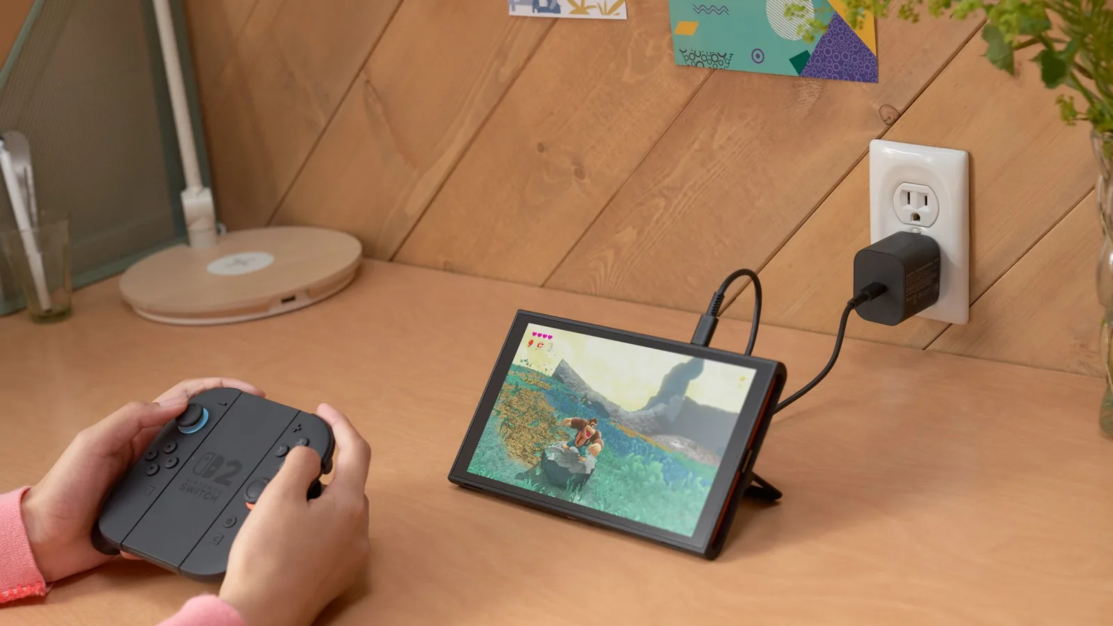 Switch 2 na stojánku se nabíjí ze zásuvky, dítě drží Joy-Con 2 v gripu